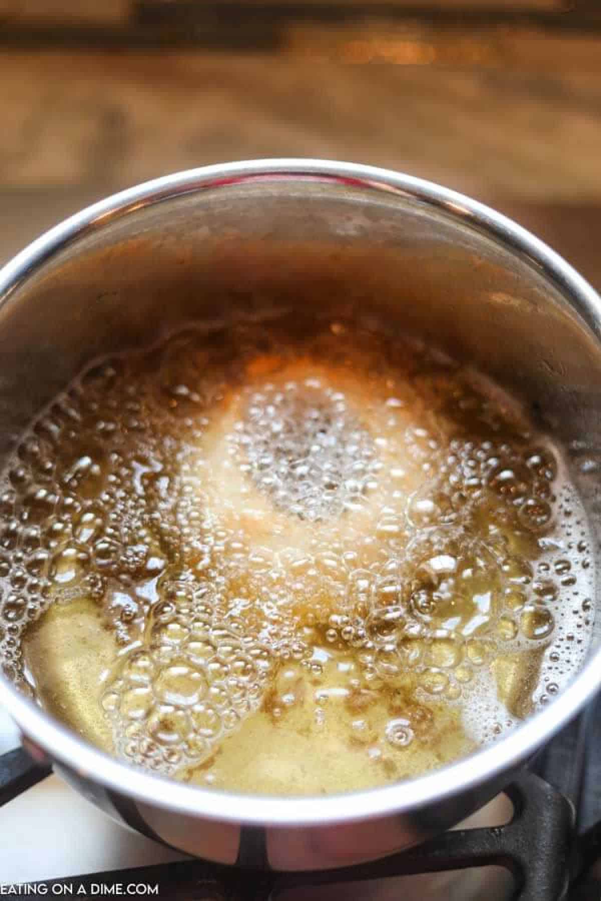 A close-up of a pot filled with bubbling caramel sauce on a stovetop, reminiscent of the golden hues found on crispy fried onion rings. The sauce is foamy and golden brown, indicating it is being heated to the boiling point.