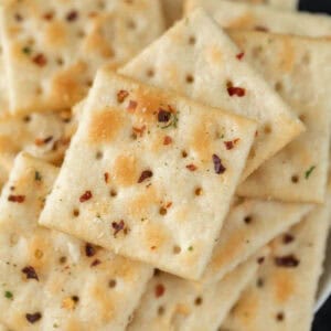 A close-up of spicy ranch crackers, square and sprinkled with herbs and red pepper flakes, stacked in a slightly overlapping arrangement.