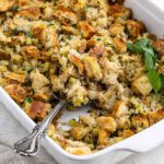 A white baking dish filled with an easy stuffing recipe made of cubed bread, celery, onions, and herbs, finished with a serving spoon and a sprig of parsley for garnish.