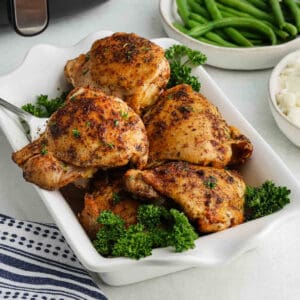 A white dish filled with four seasoned, roasted Air Fryer Chicken Thighs garnished with parsley, with bowls of green beans and mashed potatoes in the background.