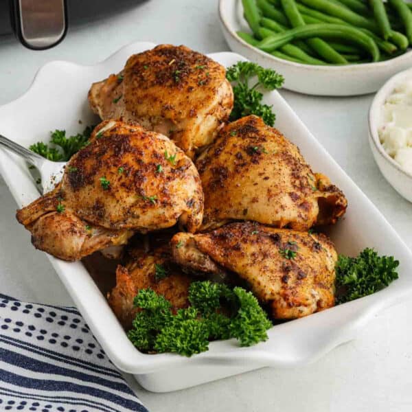 A white dish filled with four seasoned, roasted Air Fryer Chicken Thighs garnished with parsley, with bowls of green beans and mashed potatoes in the background.