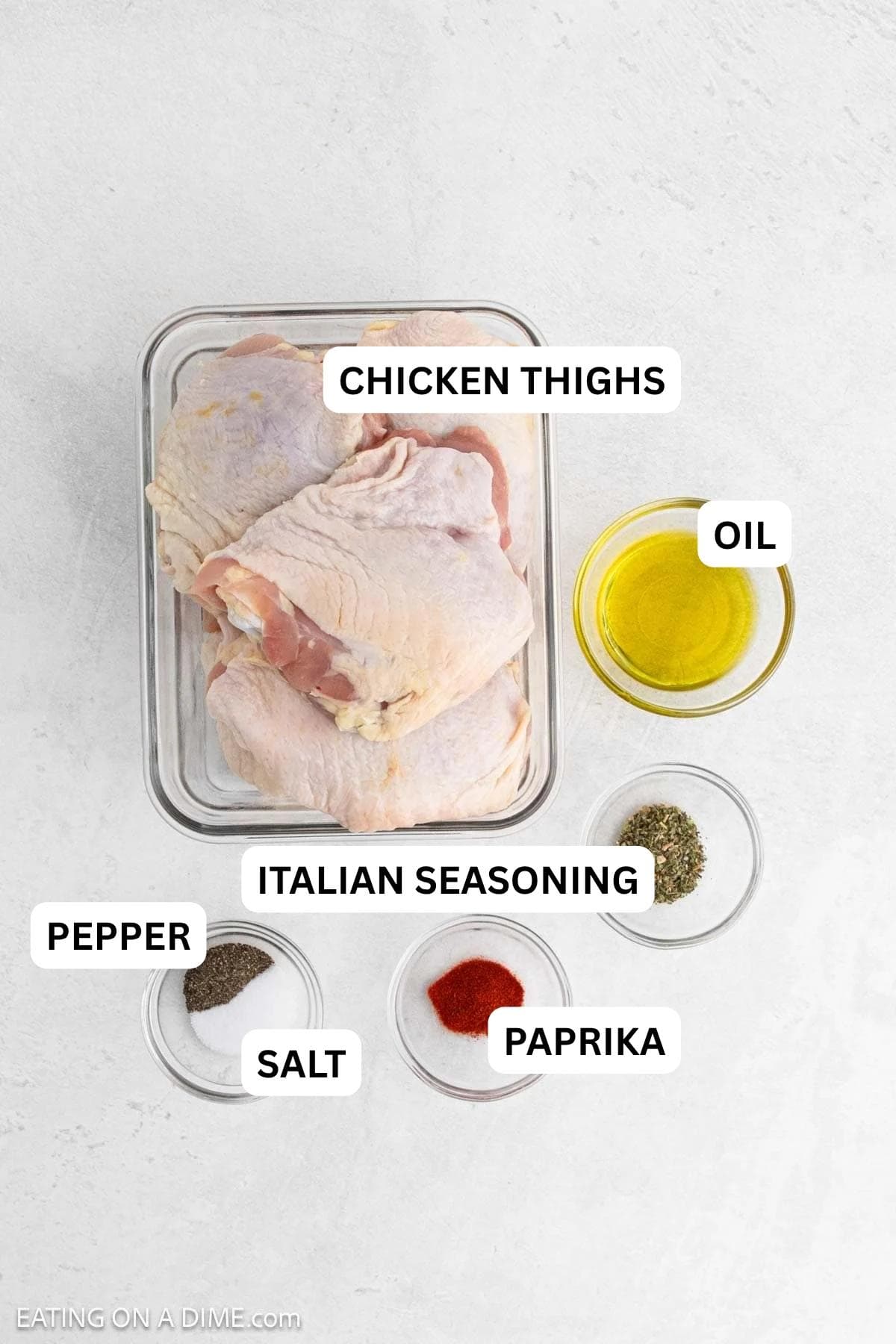Ingredients needed to make this recipe which includes chicken thighs, olive oil, dried herbs, salt, pepper, and paprika on a light surface.
