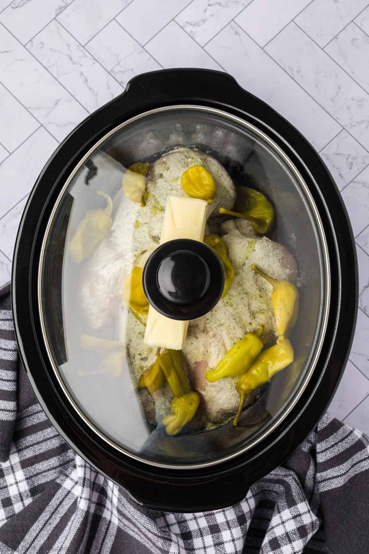 Slow cooker with a lid on top of the seasoned chicken topped with the peperoncini