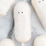A white, oval-shaped Ghost Twinkies cake pop covered in white icing with two small candy eyes, resembling a ghost, sits on a marble surface. Other similar cake pops are partially visible nearby.