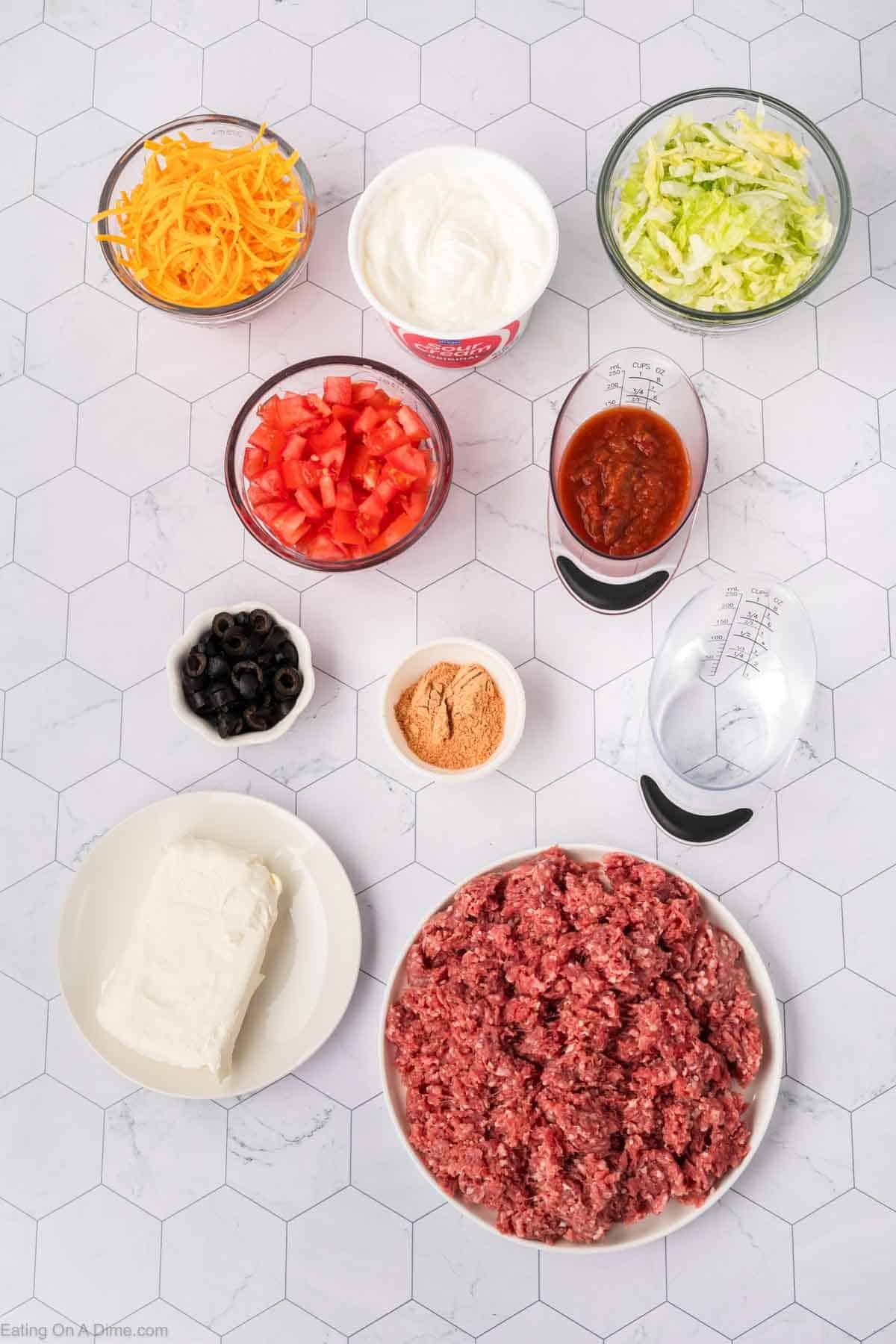 A top-down view of ground beef taco dip ingredients on a hexagonal patterned surface. Includes shredded cheese, sour cream, shredded lettuce, diced tomatoes, salsa, black olives, taco seasoning, a hearty layer of ground beef, and creamy cream cheese. Measuring cups are nearby.
