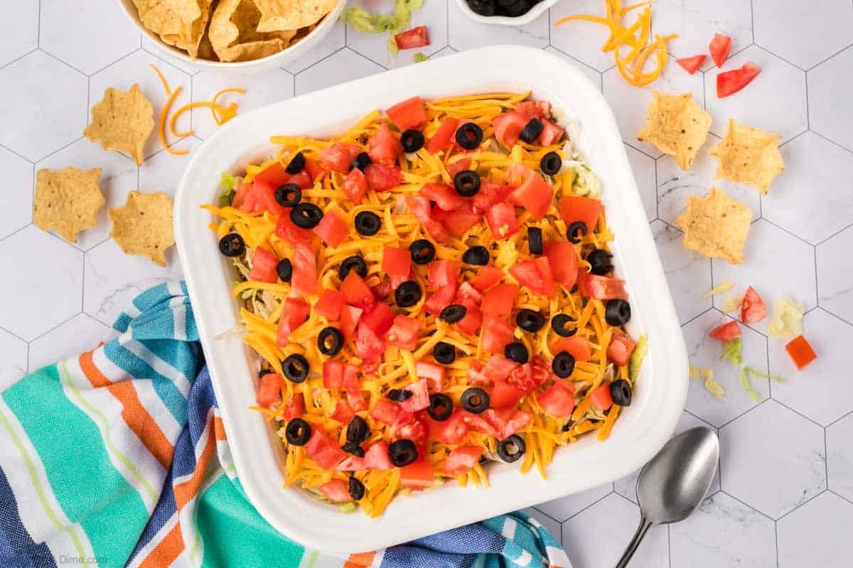 A square dish brimming with ground beef taco dip, topped with diced tomatoes, sliced black olives, and shredded cheese. Surrounding the dish are tortilla chips and a colorful striped cloth. A spoon rests beside the dish on a hexagonal-tiled surface.