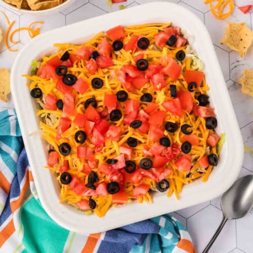 A square dish of layered ground beef taco dip is topped with diced tomatoes, shredded cheddar cheese, and sliced black olives. A colorful striped towel and spoon sit beside the dish, with tortilla chips scattered nearby on a geometric-patterned surface.