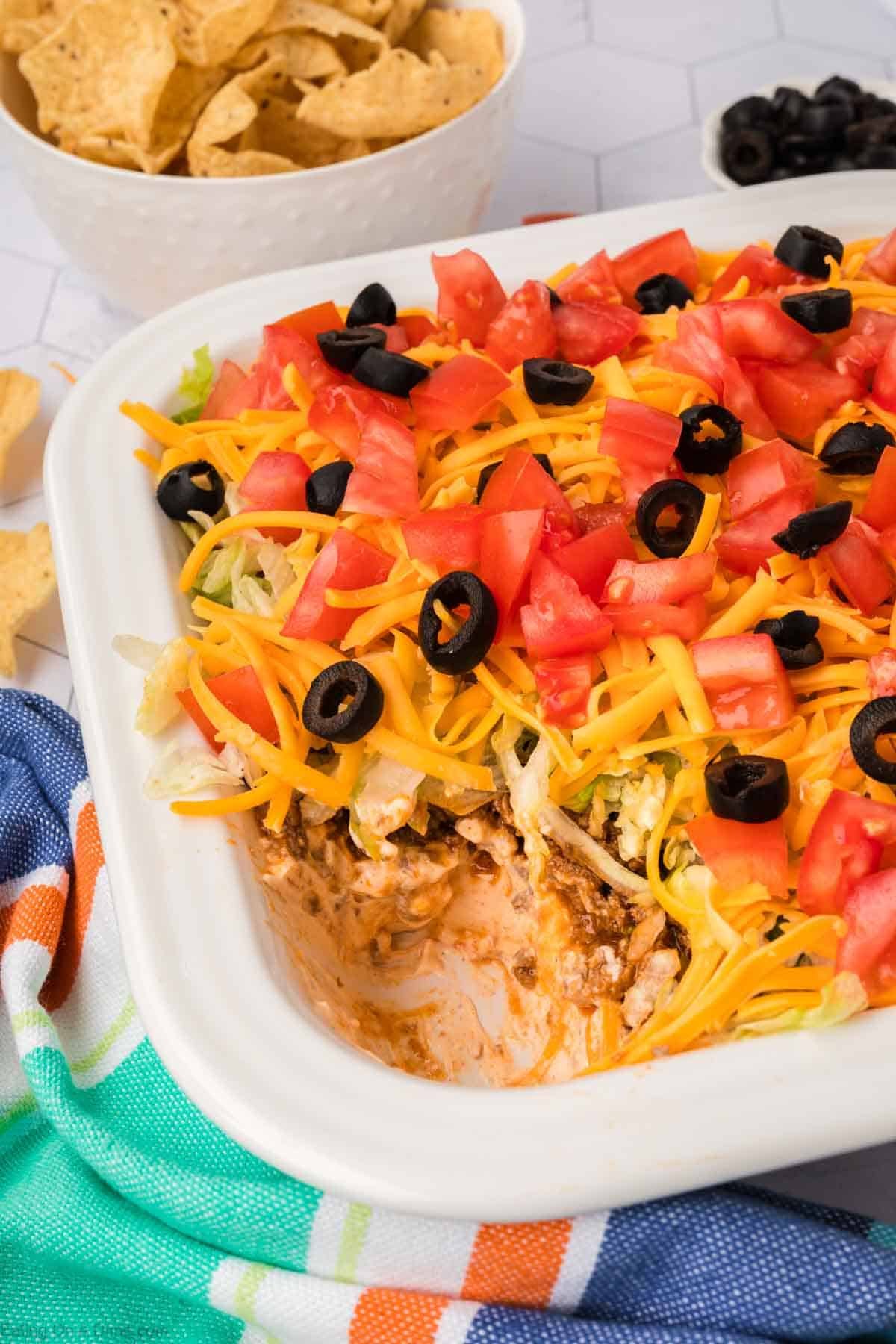 A rectangular dish filled with layered ground beef taco dip is topped with chopped tomatoes, shredded cheese, and sliced black olives. Some of the savory dip is scooped out, revealing layers underneath. A bowl of tortilla chips waits enticingly in the background.