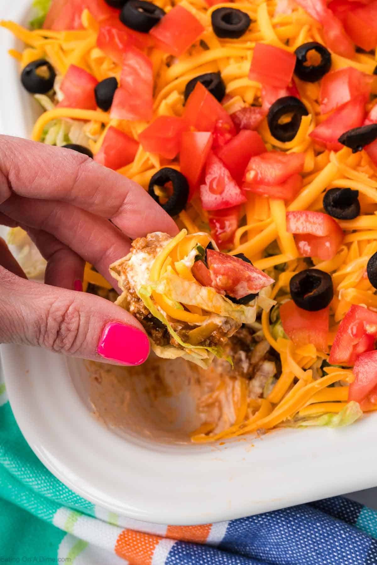 A hand with pink nail polish holds a tortilla chip loaded with layers of cheese, tomato, lettuce, sour cream, and savory ground beef taco dip. The background reveals a dish brimming with similar toppings like sliced black olives and chopped tomatoes on a vibrant cloth.