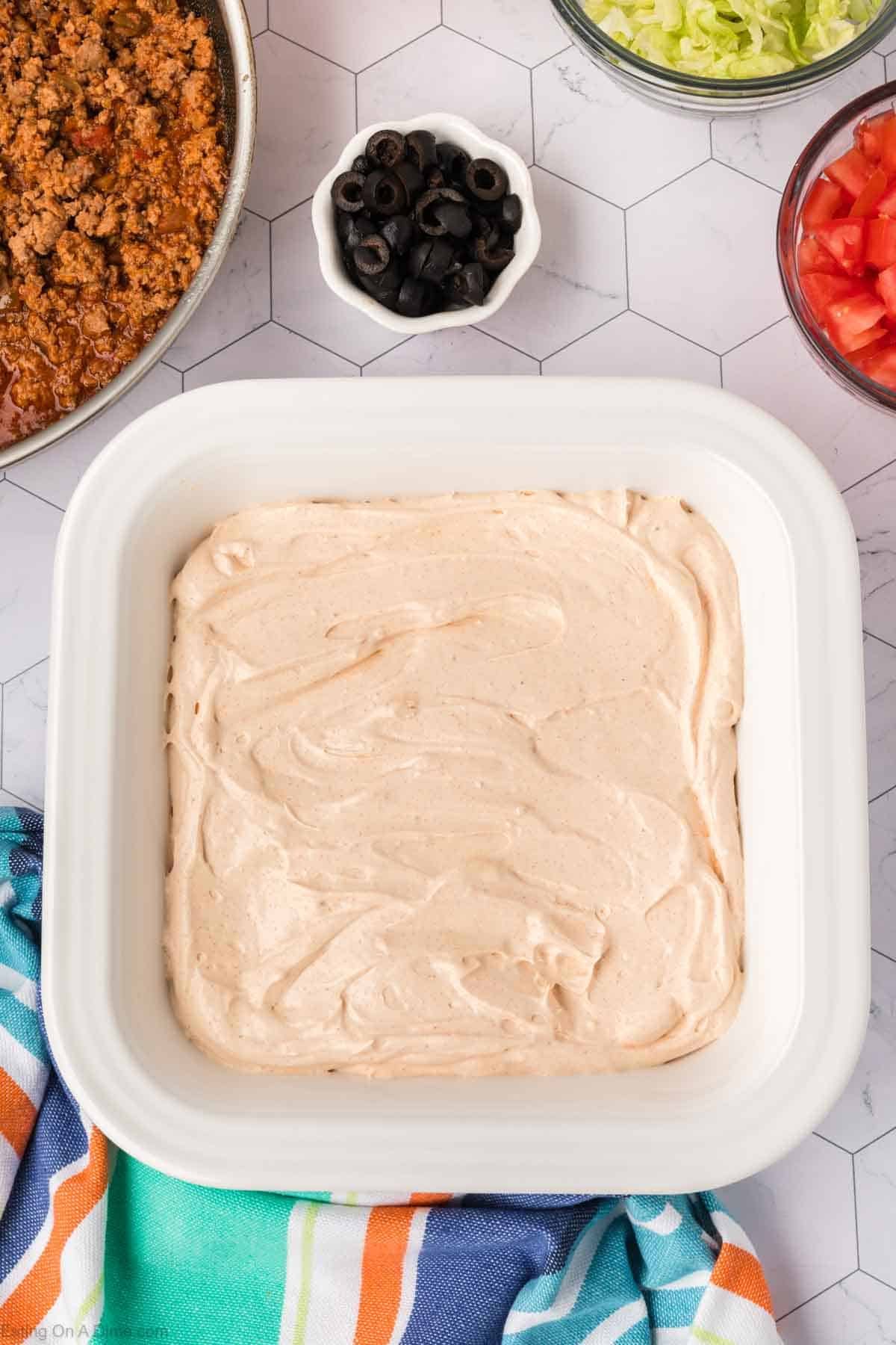 A square dish of creamy ground beef taco dip sits surrounded by toppings: sliced black olives, chopped lettuce, and diced tomatoes. A colorful striped cloth is casually placed beside the dish. The background features a hexagonal pattern.
