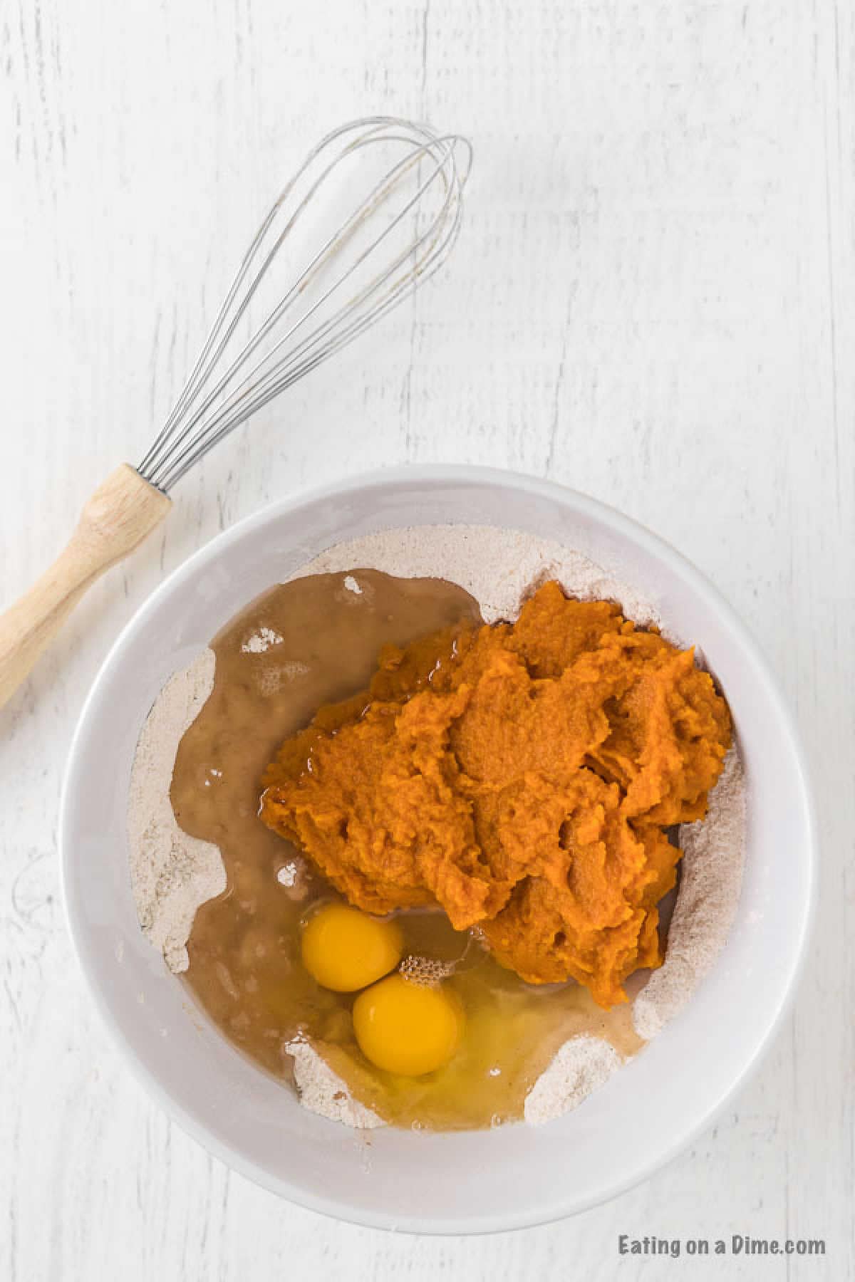 A white bowl contains a mixture of flour, two cracked eggs, pumpkin puree, and a liquid ingredientโa perfect start to a pumpkin bread recipe. A whisk with a wooden handle rests beside the bowl on a white surface.