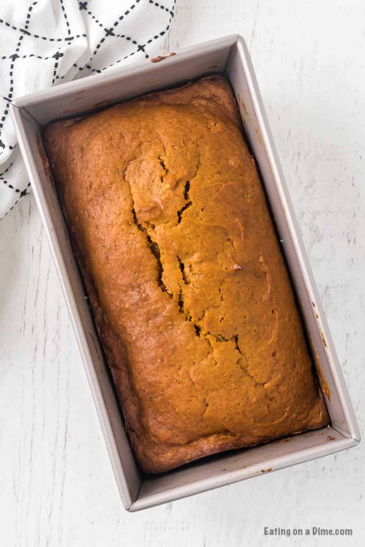 This is the BEST pumpkin bread recipe. It is easy to make and this Pumpkin Bread Recipe tastes amazing! Try this recipe for Pumpkin Bread. A loaf of freshly baked pumpkin bread rests in a rectangular pan on a white surface, as if straight from a cherished pumpkin bread recipe. A checkered black and white cloth accents the scene's rustic charm, while the golden-brown crust boasts a delightful crack on top.