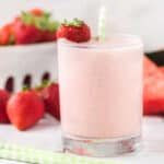 A glass of creamy pink Strawberry Watermelon Smoothie with a green-and-white striped straw, garnished with a whole strawberry. Fresh strawberries are scattered in the background.