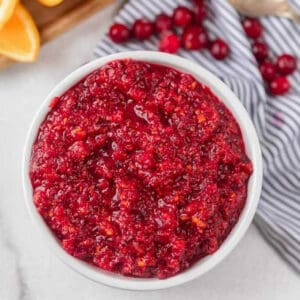 A white bowl filled with chunky Fresh Cranberry Relish sits on a marble surface, with a few fresh cranberries and a striped napkin nearby.