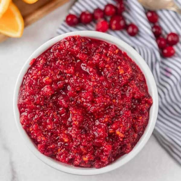 A white bowl filled with chunky Fresh Cranberry Relish sits on a marble surface, with a few fresh cranberries and a striped napkin nearby.