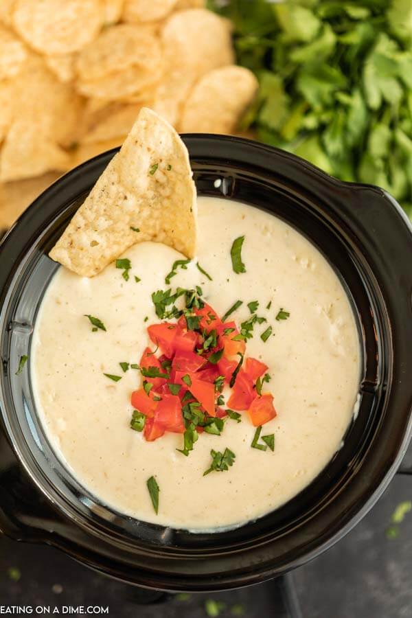 White Cheese Dip in a small crock pot topped with diced tomatoes, cilantro and a chip dipped into it with more chips behind it 
