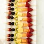 A row of colorful fruit kabobs—wooden skewers with grapes, blackberries, kiwi, pineapple, cantaloupe, and strawberries—are neatly arranged on a rectangular white platter.