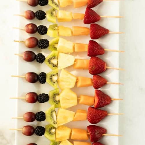 A row of colorful fruit kabobs—wooden skewers with grapes, blackberries, kiwi, pineapple, cantaloupe, and strawberries—are neatly arranged on a rectangular white platter.