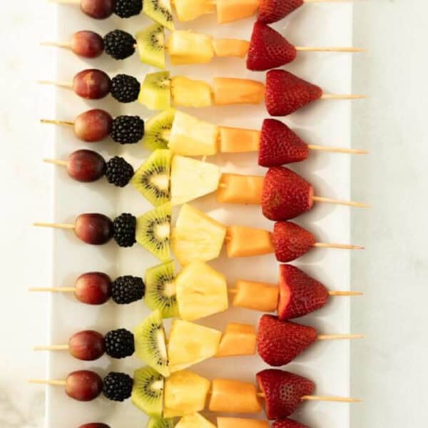 A row of colorful fruit kabobs—wooden skewers with grapes, blackberries, kiwi, pineapple, cantaloupe, and strawberries—are neatly arranged on a rectangular white platter.