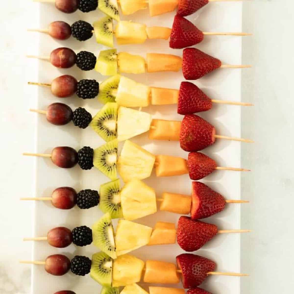 A row of colorful fruit kabobs—wooden skewers with grapes, blackberries, kiwi, pineapple, cantaloupe, and strawberries—are neatly arranged on a rectangular white platter.