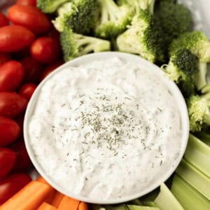 A bowl of creamy homemade ranch dip garnished with herbs, surrounded by fresh vegetables including cherry tomatoes, broccoli florets, celery sticks, and carrot sticks.