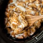A wooden spoon lifts a gooey portion of Crock Pot Cinnamon Roll Casserole, drizzled with white icing, above a slow cooker brimming with more of the decadent dessert.