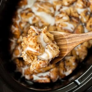 A wooden spoon lifts a gooey portion of Crock Pot Cinnamon Roll Casserole, drizzled with white icing, above a slow cooker brimming with more of the decadent dessert.