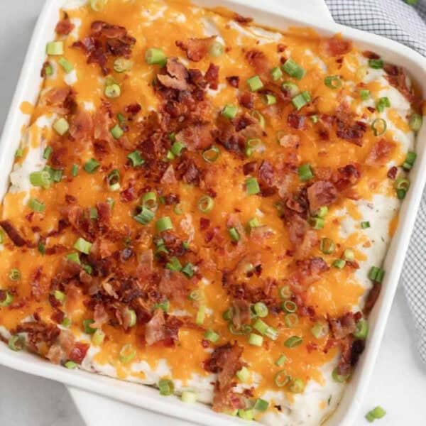 A baked casserole of loaded mashed potatoes topped with melted cheddar cheese, crispy bacon bits, and chopped green onions, served in a white square dish.