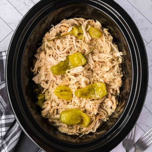 Shredded Crock Pot Mississippi Chicken topped with whole pepperoncini peppers in a black slow cooker, set on a white marble surface next to a black and white checkered napkin and silverware.