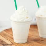A vanilla milkshake in a clear plastic cup topped with whipped cream, with a green straw, placed on a wooden surface—just like the Starbucks vanilla bean frappuccino recipe. Another similar milkshake is partially visible in the background.