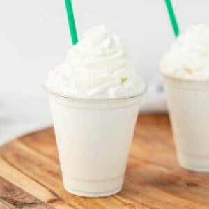A vanilla milkshake in a clear plastic cup topped with whipped cream, with a green straw, placed on a wooden surface—just like the Starbucks vanilla bean frappuccino recipe. Another similar milkshake is partially visible in the background.