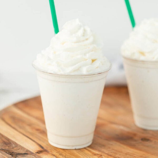 A vanilla milkshake in a clear plastic cup topped with whipped cream, with a green straw, placed on a wooden surface—just like the Starbucks vanilla bean frappuccino recipe. Another similar milkshake is partially visible in the background.