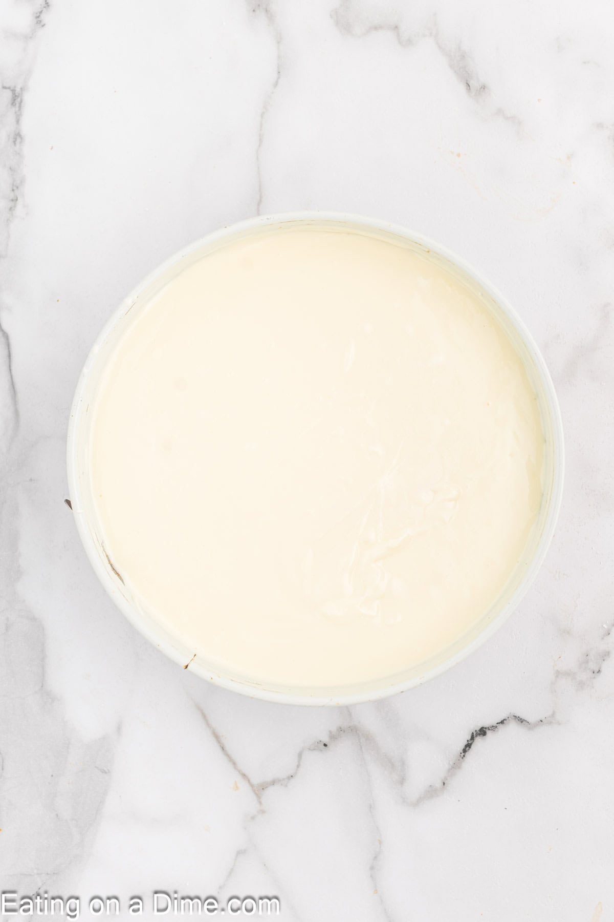 A top-down view of a white bowl containing a smooth, creamy white substance resembling yogurt or a similar dairy product, placed on a white marble countertop. The surface of the creamy substance is even and glossy, evoking the rich texture of Pecan Pie Cheesecake filling.