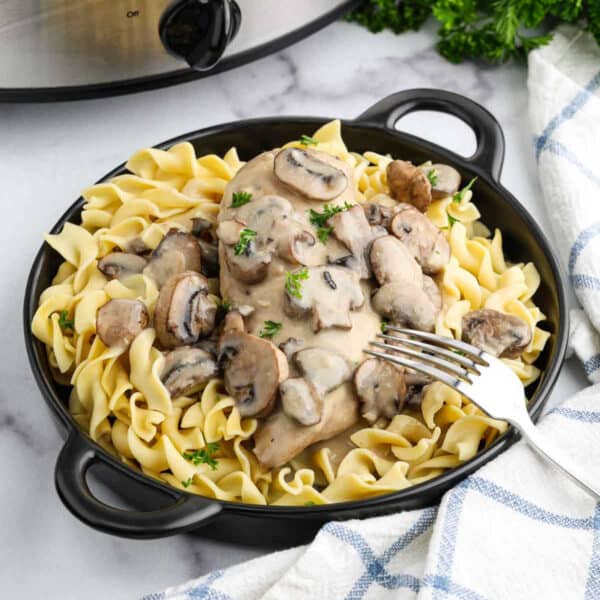 A black bowl filled with egg noodles topped with creamy crock pot chicken marsala and sliced mushrooms, garnished with parsley. A fork rests on the side and a slow cooker is in the background.