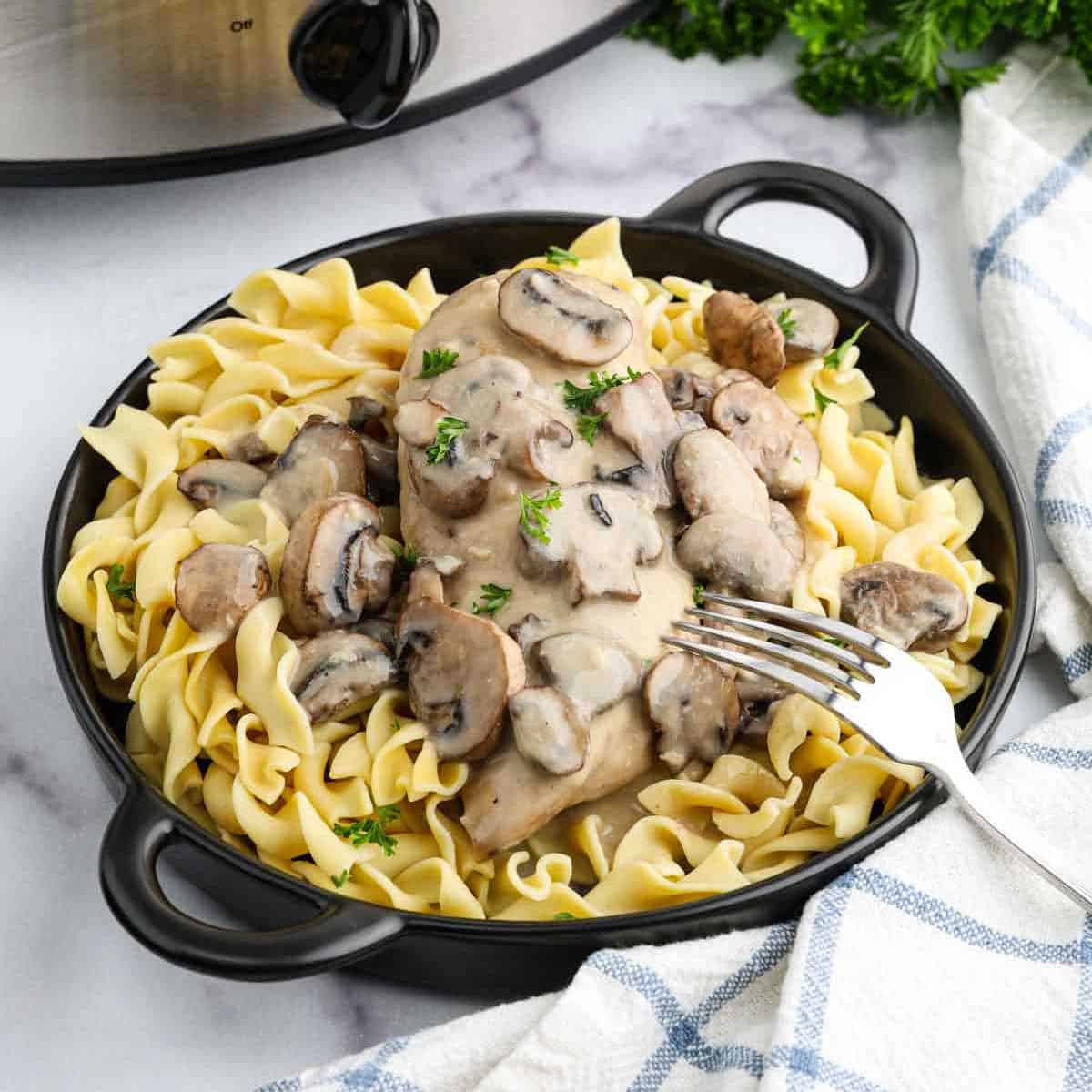 A black bowl filled with egg noodles topped with creamy crock pot chicken marsala and sliced mushrooms, garnished with parsley. A fork rests on the side and a slow cooker is in the background.