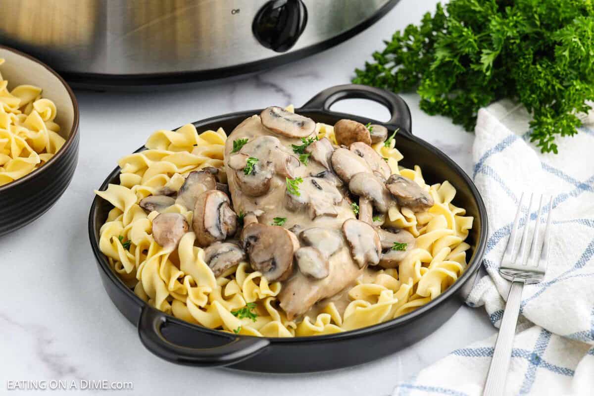 A black bowl filled with egg noodles topped with creamy crock pot chicken marsala, garnished with parsley. Fresh parsley, a fork, a napkin, and a slow cooker are in the background.
