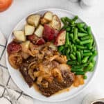 A white plate filled with tender Instant Pot steak pot roast topped with onions, boiled red potatoes, and cooked green beans, placed on a light surface next to a napkin.