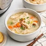 A bowl of Instant Pot Chicken Noodle Soup with shredded chicken, carrots, celery, and noodles, garnished with parsley. A cracker and a fork are nearby, and a pressure cooker is visible in the background.