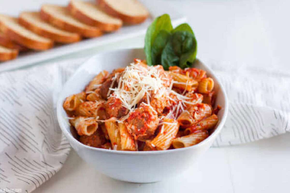 This Crock pot Baked Pasta with Meatballs recipe is tasty and takes minutes to toss in your crockpot. Slow cooker pasta bake with meatballs is so easy. A bowl of Crockpot Pasta and Meatballs features rigatoni topped with meatballs, tomato sauce, grated cheese, and fresh spinach leaves. Slices of bread are visible in the background.