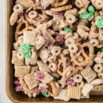 A close-up of a baking tray filled with Christmas Chex Mix, featuring cereal squares, pretzels, round oat cereal, and red and green candy-coated chocolates, all coated in a white, sugary glaze.