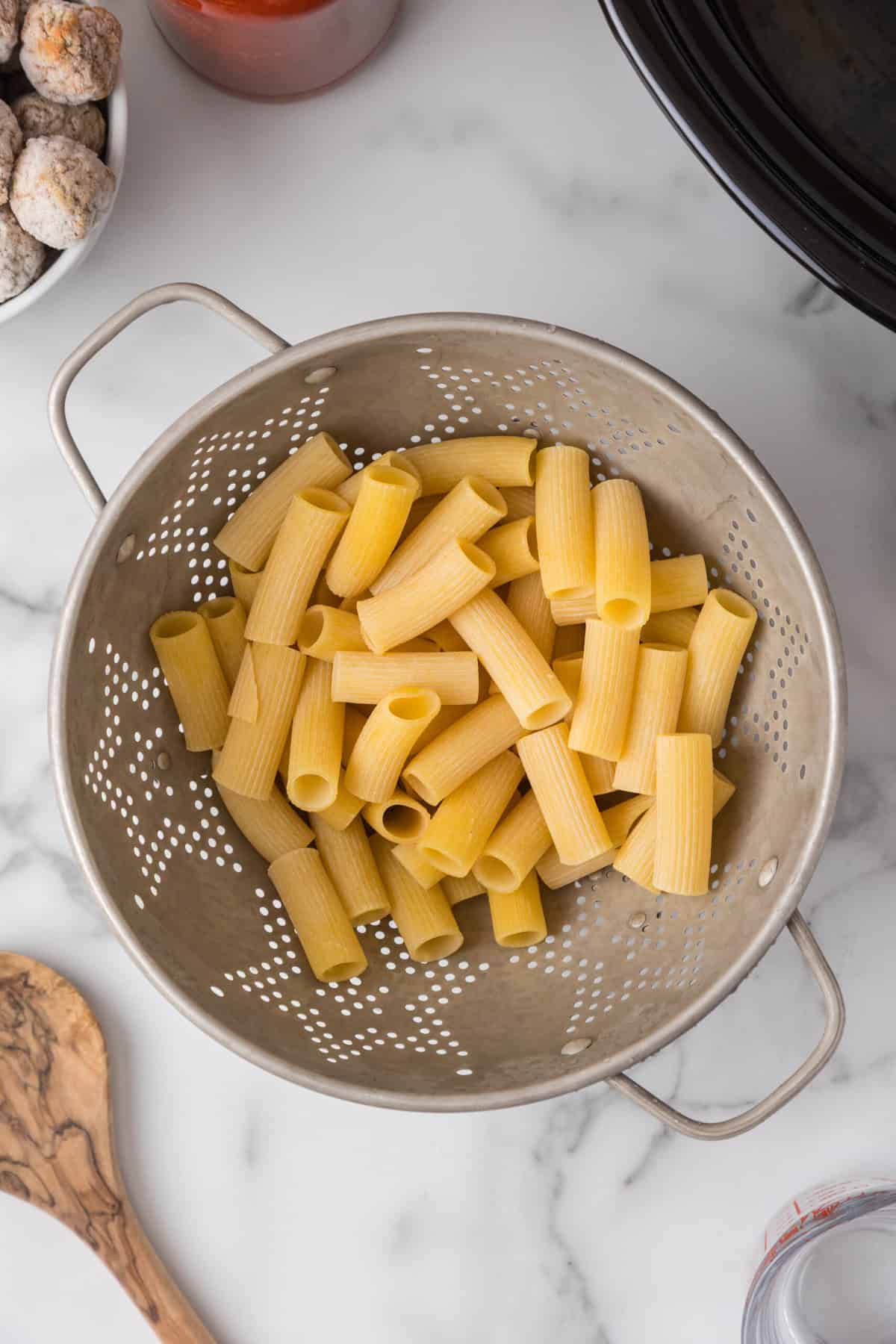 Uncooked rigatoni pasta in a metal colander on a marble countertop, with a wooden spoon, a bowl of meatballs, and a black pot partially visible—perfect ingredients for an easy Crockpot Pasta and Meatballs dinner.