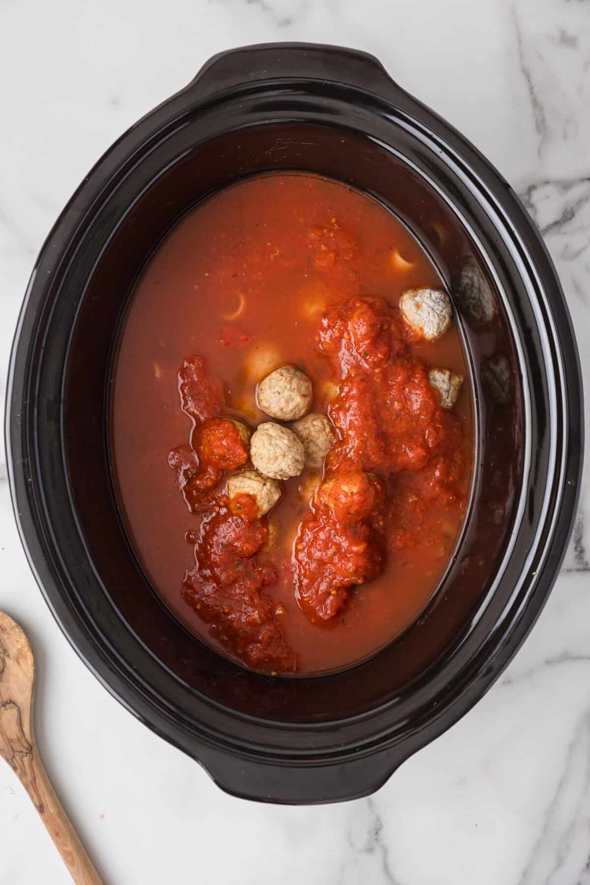 A black slow cooker filled with tomato sauce and several meatballs, perfect for Crockpot Pasta and Meatballs, sits on a white marble surface. A wooden spoon is partially visible on the left side of the image.