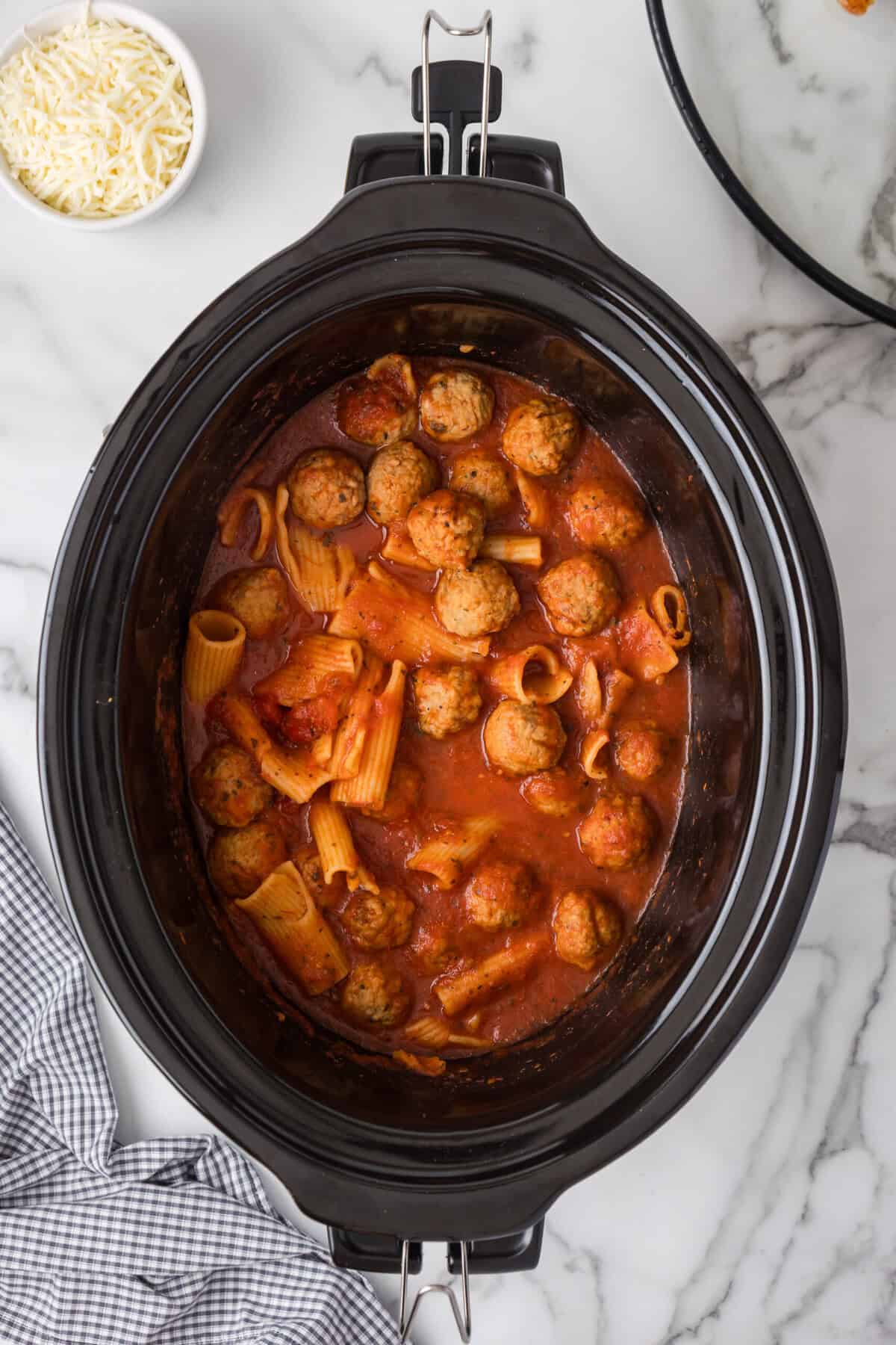 A Crockpot Pasta and Meatballs dish with rigatoni, meatballs, and tomato sauce simmers on a marble countertop. Nearby, a bowl of shredded cheese and a checkered cloth await.