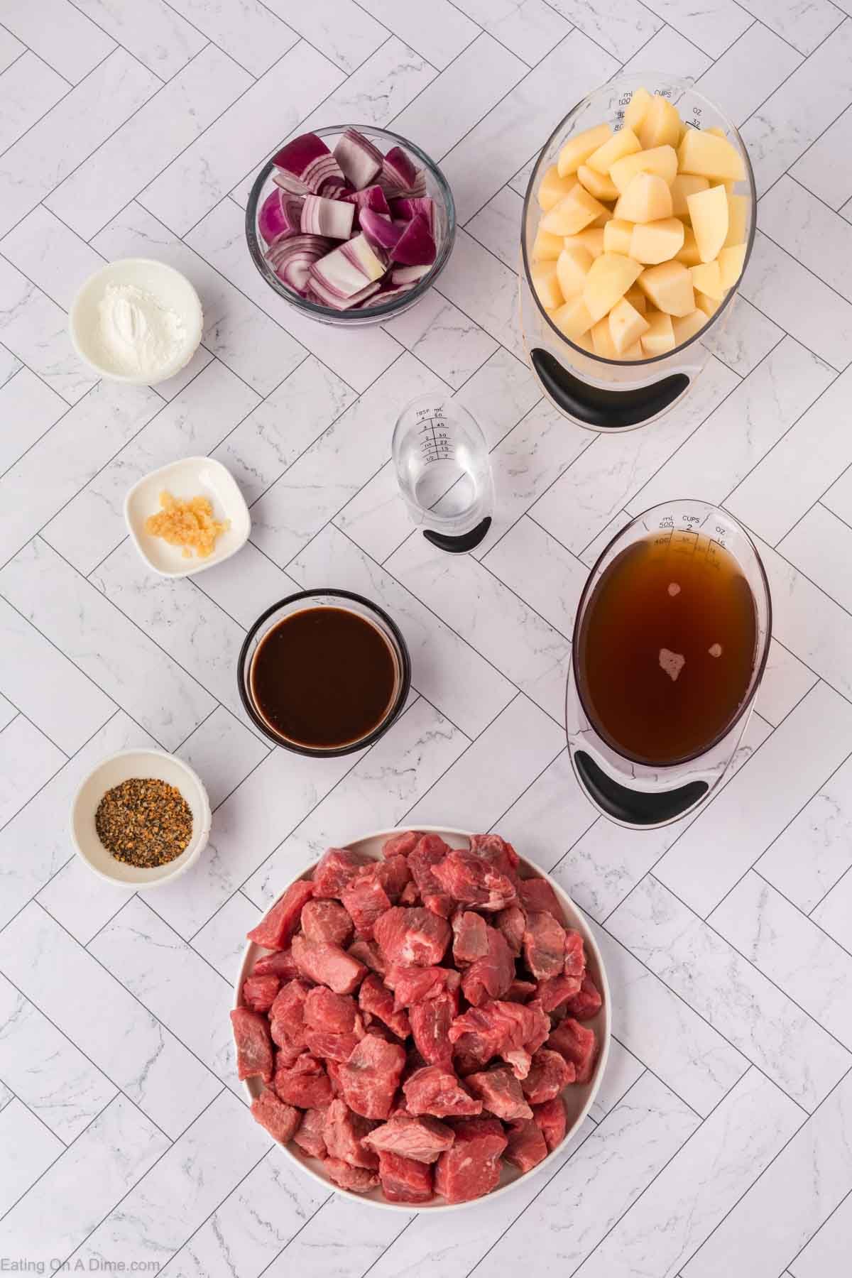 Overhead view of raw beef chunks, diced potatoes, chopped red onions, beef broth, minced garlic, flour, seasoning mix, and Worcestershire sauce arranged in separate bowls—perfect for a crock pot steak and potato stew recipe.