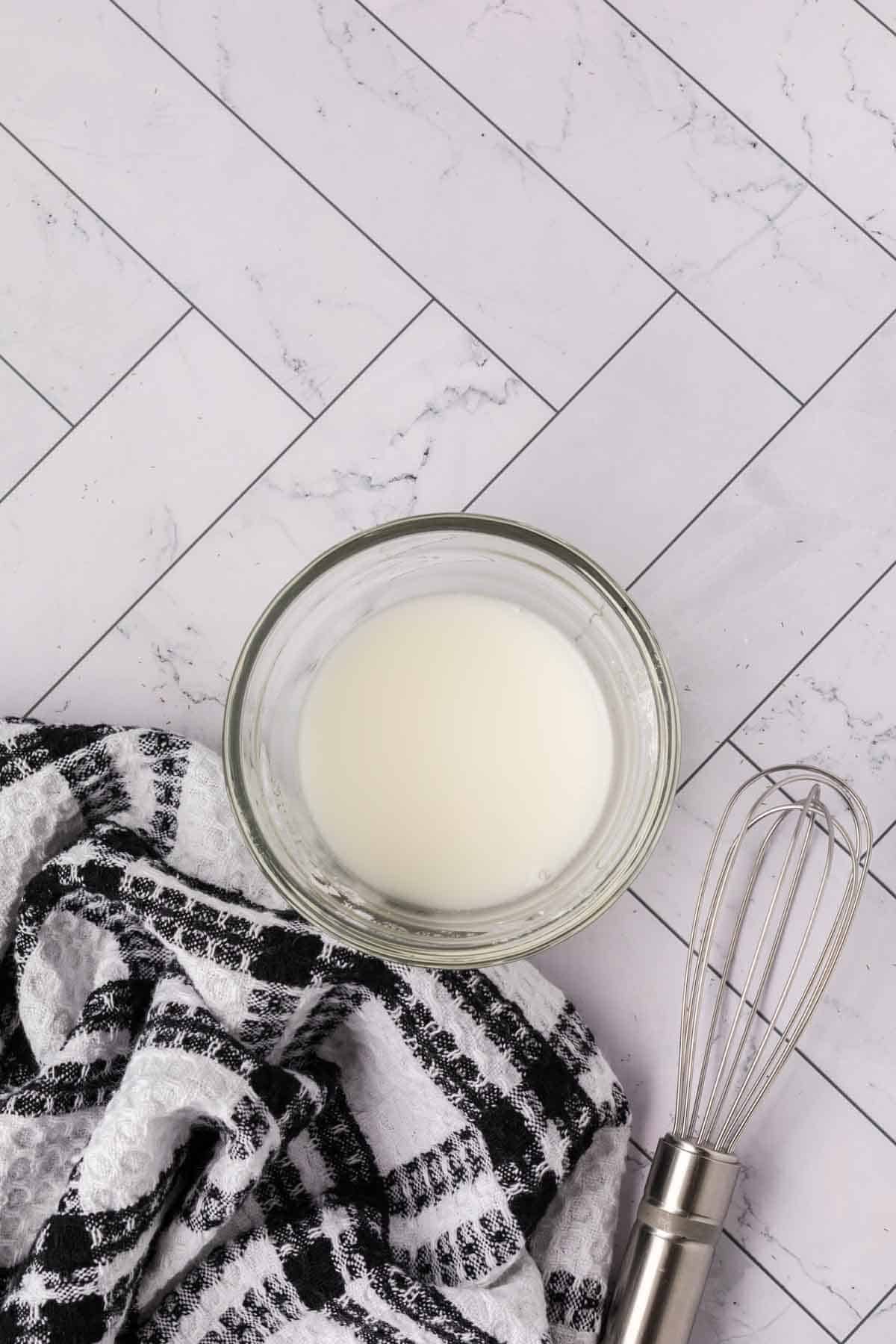 A glass bowl filled with a white liquid sits on a white tiled surface next to a black and white checkered kitchen towel and a metal whisk, ready for preparing ingredients for your crock pot steak and potato stew recipe.