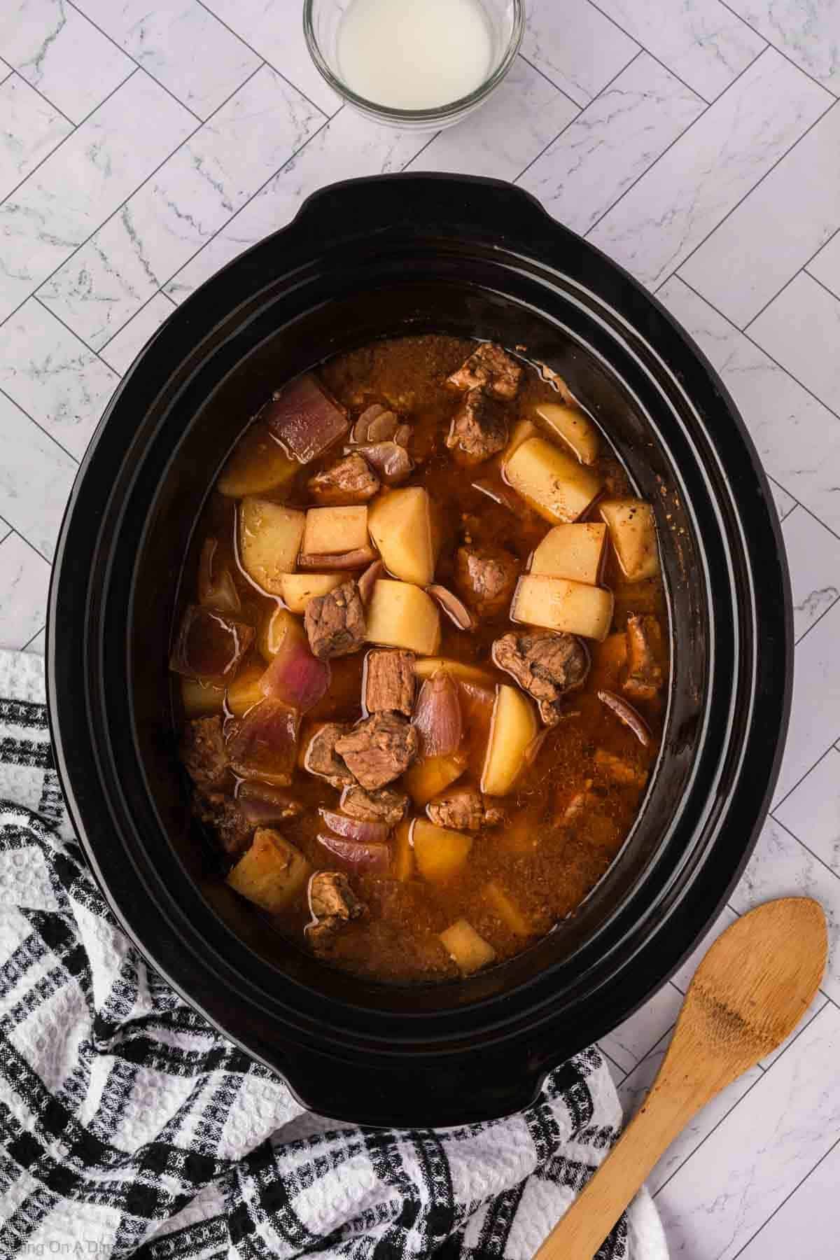 A black slow cooker filled with crock pot steak and potato stew sits on a white tiled surface. Chunks of beef, potatoes, and onions are visible. Nearby are a wooden spoon, a glass of milk, and a black-and-white checkered towel.