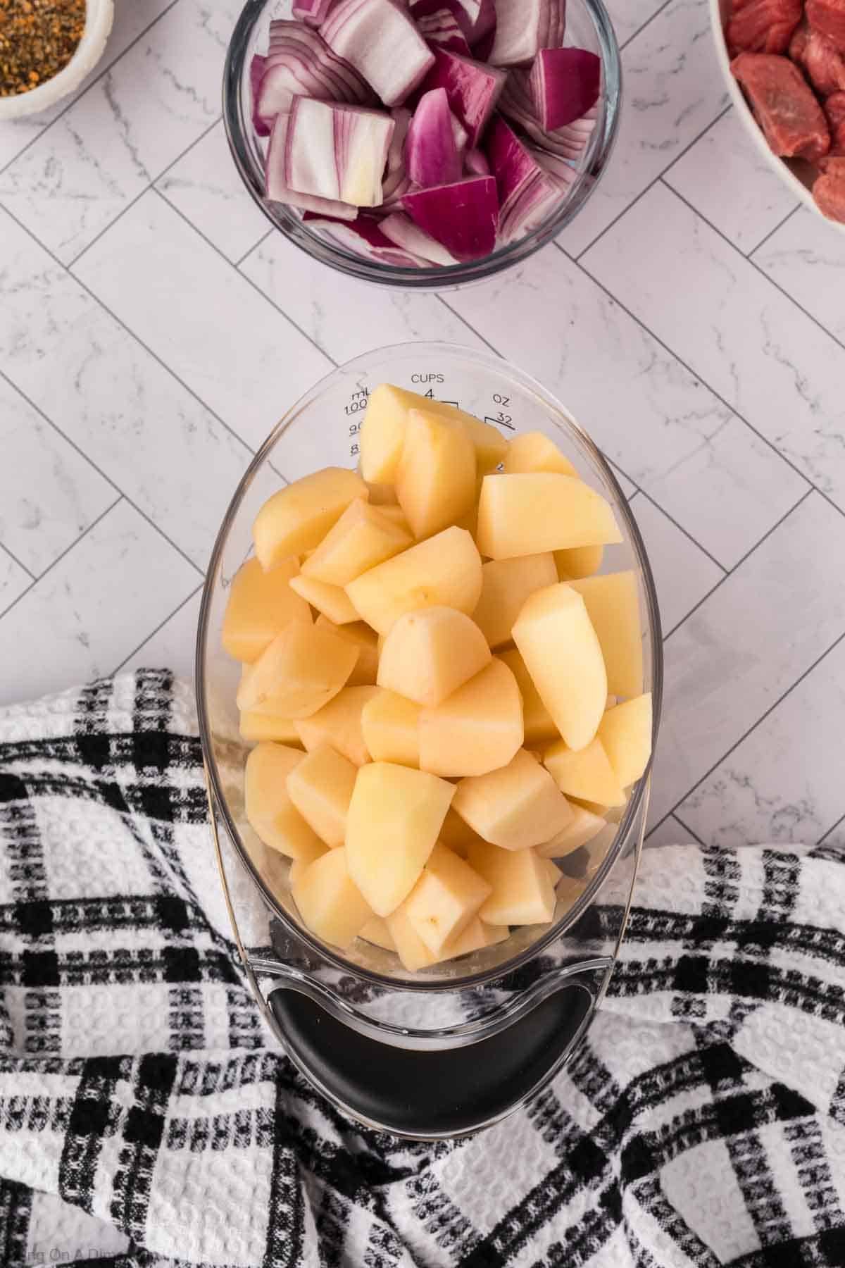 A clear glass bowl filled with peeled and chopped potatoes sits on a black and white checkered towel, surrounded by bowls of chopped red onions and chunks of raw meat—perfect for prepping a crock pot steak and potato stew recipe.