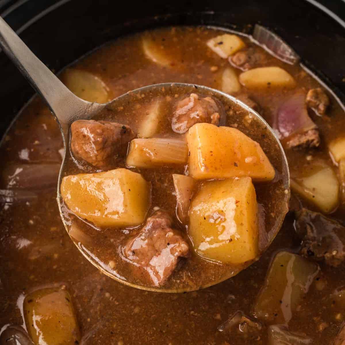 A ladle filled with hearty crock pot steak and potato stew recipe—tender beef, potatoes, and carrots in a rich brown broth—held above a simmering pot brimming with more savory stew.
