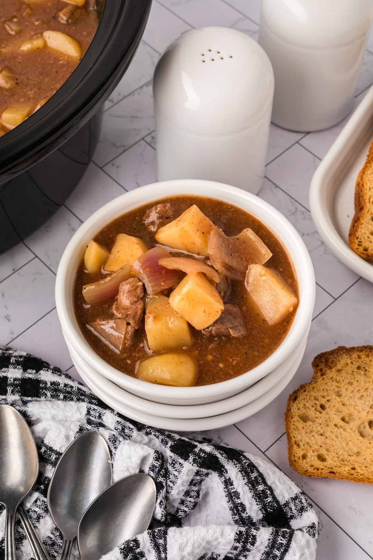 A bowl of crock pot steak and potato stew sits on a stacked white plate, next to toast, a black-and-white towel, and several spoons. A slow cooker and salt and pepper shakers are in the background.
