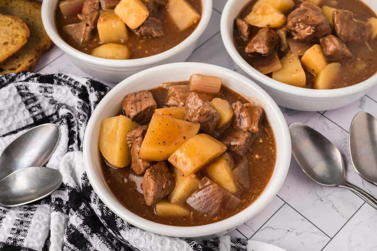 Three bowls of beef stew with chunks of beef, potatoes, and carrots in brown broth—just like a classic crock pot steak and potato stew recipe—are placed on a white tiled surface with spoons, toasted bread, and a black-and-white cloth napkin nearby.